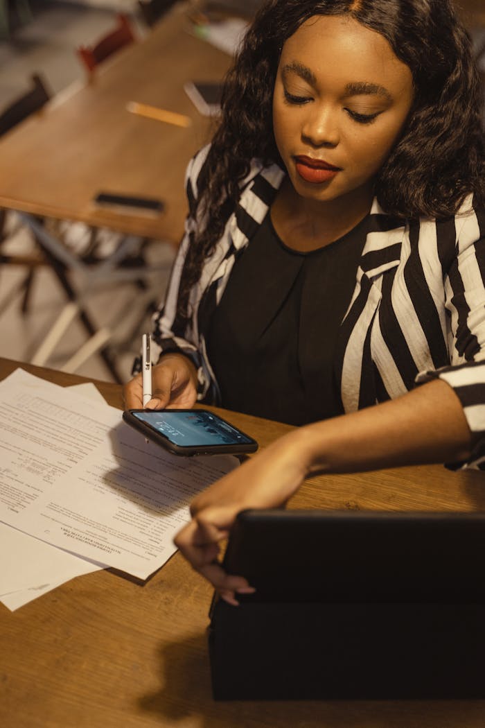 A businesswoman with curly hair reviews documents while using her smartphone at a table.