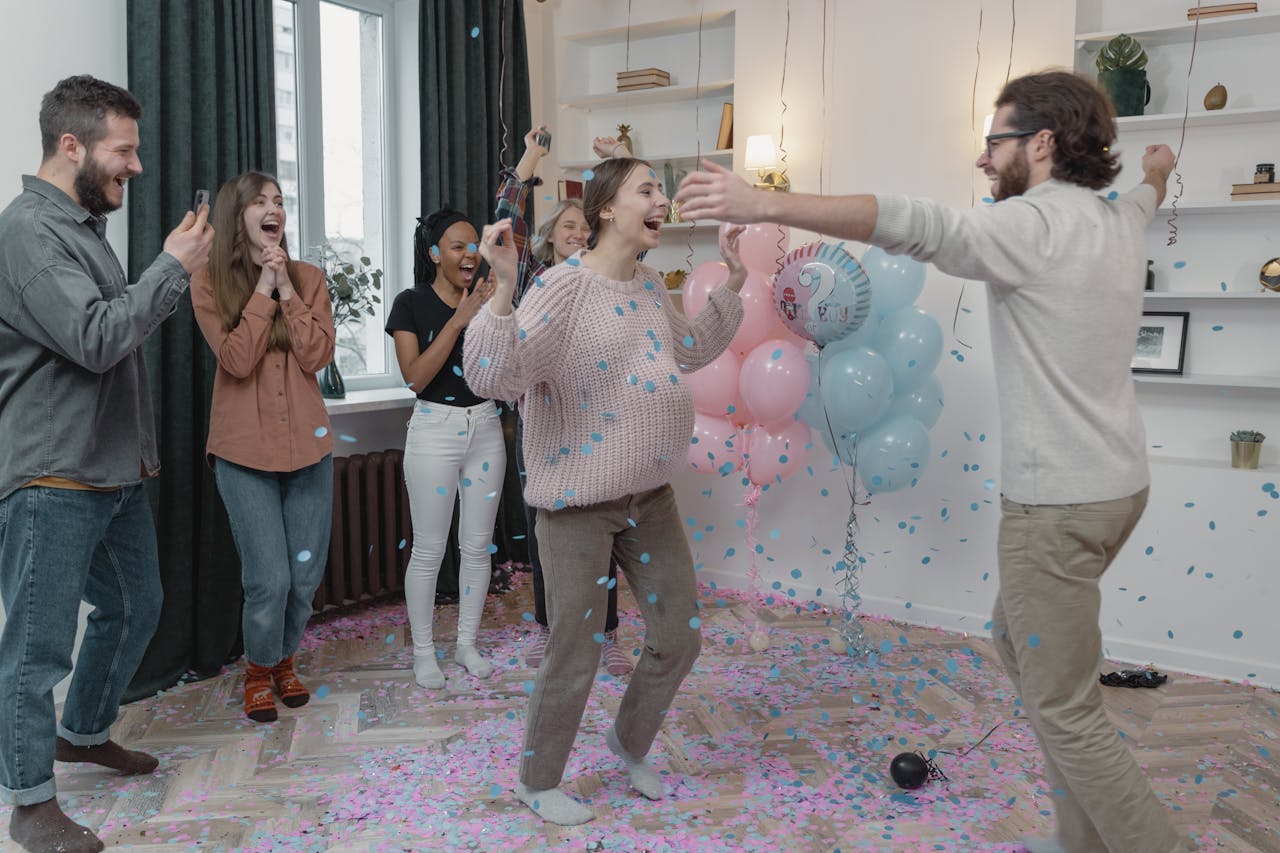 Happy friends celebrate a gender reveal party with confetti and balloons indoors.